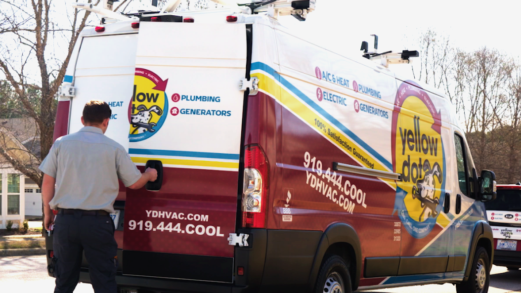 A technician opens the back door of a Yellow Dot service van parked outdoors. The van displays HVAC, plumbing, electric, and generator services and contact details.