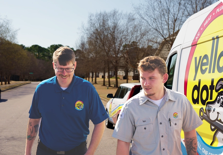 Two men in company uniforms stand outdoors near branded service vans on a residential street, with trees and houses in the background.
